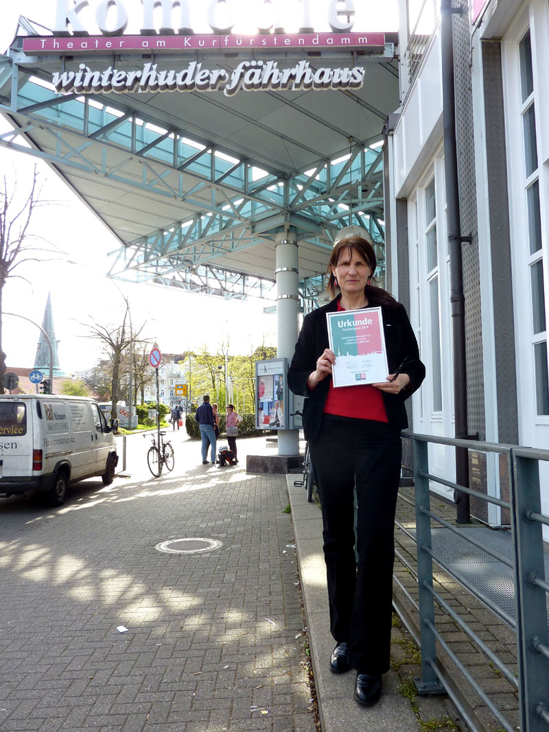 Gisela Baudy mit Urkunde und Elbbrücken-Plakat vor der Komödie des Winterhuder Fährhauses. (Foto Chris Baudy)